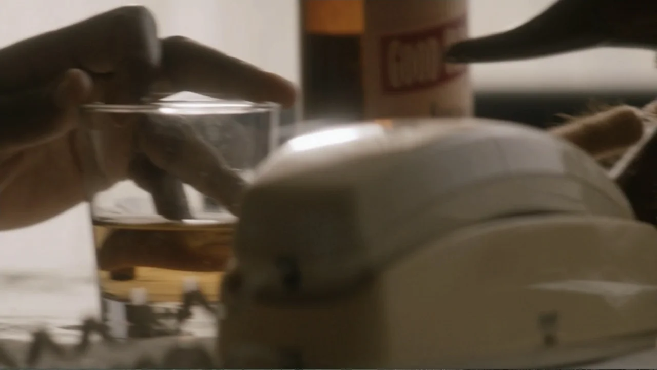 Nightingale (2014) movie still: closeup — A cluttered desk surface features a beige rotary phone and a glass of amber liquid. A hand rests near the…