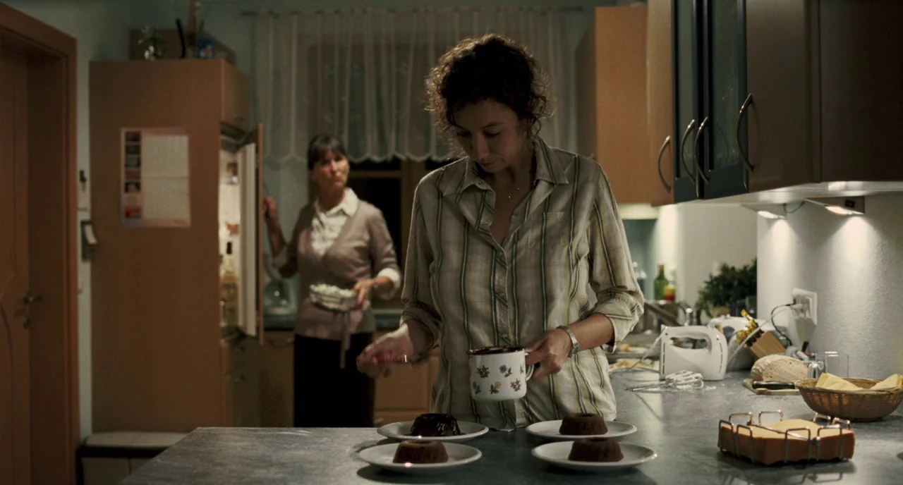 Revanche (2008) movie still: medium — A dimly lit home kitchen with laminate counters, overhead cupboards, and a refrigerator. A woman in a striped…