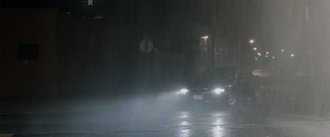 A Walk Among the Tombstones (2014) movie still: wide — A dark city street at night is saturated with heavy falling rain. A black sedan approaches…
