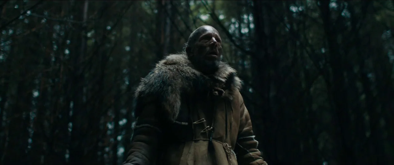 Out of Darkness (2022) movie still: medium — A dense, dark forest with blurred tree trunks filling the frame background. A man stands in the…