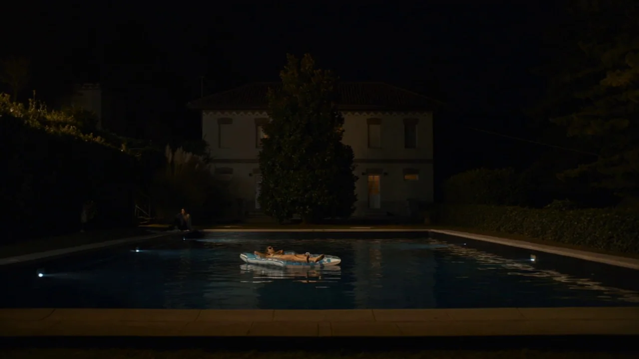 Amanda (2022) movie still: wide — A nighttime view of a rectangular pool situated in front of a multi-story residence. A person floats on a blue…