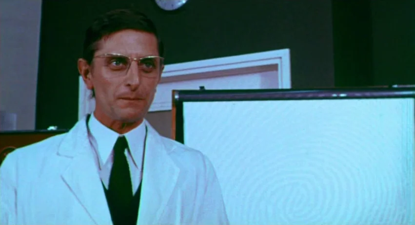A Lizard In A Woman’s Skin (1971) movie still: medium — A man in a white lab coat stands before a white board in a dark room. He is positioned in…