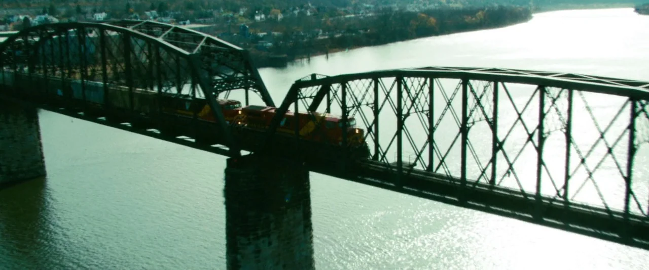 Unstoppable (2010) movie still: wide — A metal truss bridge spans a wide river under a bright overcast sky. A large freight train travels across the…