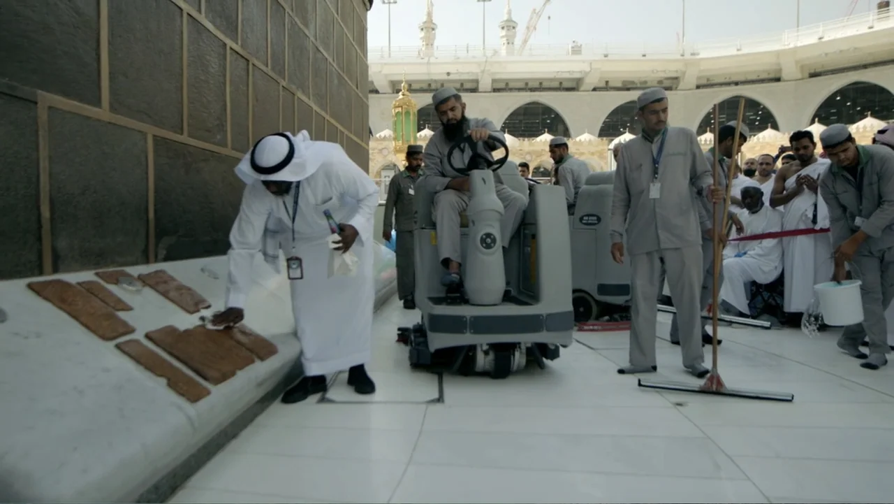 One Day in the Haram (2017) movie still: fullbody — An open white marble courtyard of a large mosque with arched architectural features. Several men…