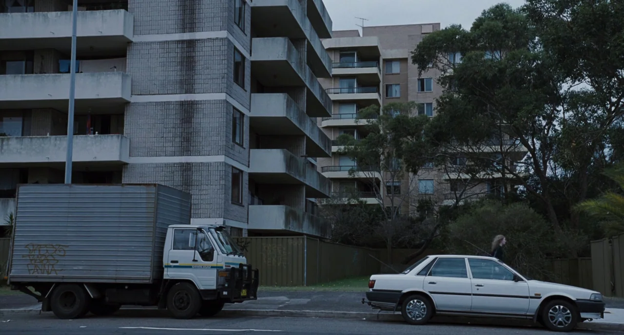 Sleeping Beauty (2011) movie still: wide — A suburban street scene features a grey concrete apartment complex and parked vehicles. A white sedan and…