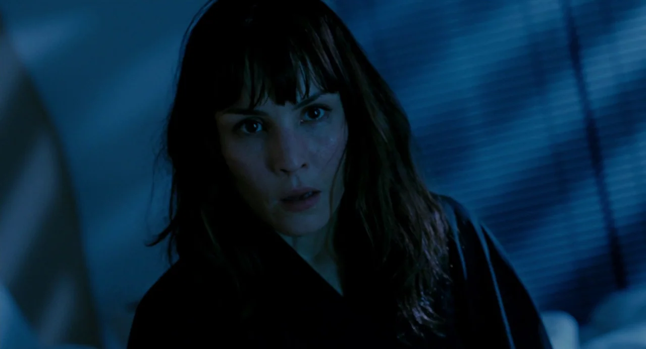 Passion (2012) movie still: closeup — A dark space features a woman in the foreground with window blinds visible behind. The woman stares forward…