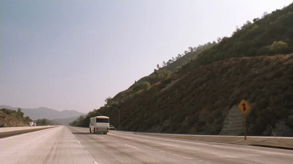 Year Of The Dog (2007) movie still: wide — An empty multi-lane concrete highway runs parallel to a large, dry, hilly landscape. A single white bus…