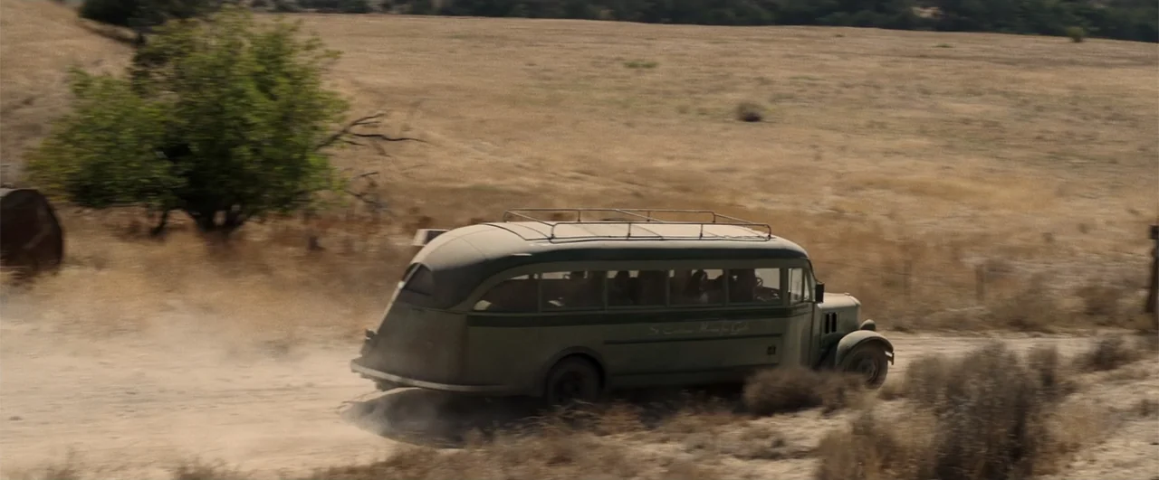 Annabelle: Creation (2017) movie still: wide — A vintage olive-drab bus travels along a dry, unpaved road through a sparse field. The bus kicks up a…