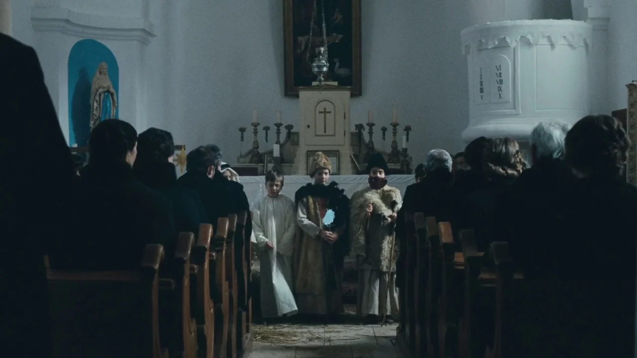 Childhood of a Leader (2015) movie still: medium — A rural church interior features white walls, dark wooden pews, and religious altar icons. Three…