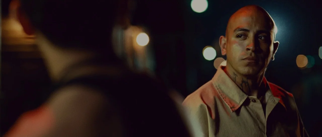 Sin Nombre (2009) movie still: medium — A dark urban environment with distant street lights creating circular bokeh in the background. A man stands…