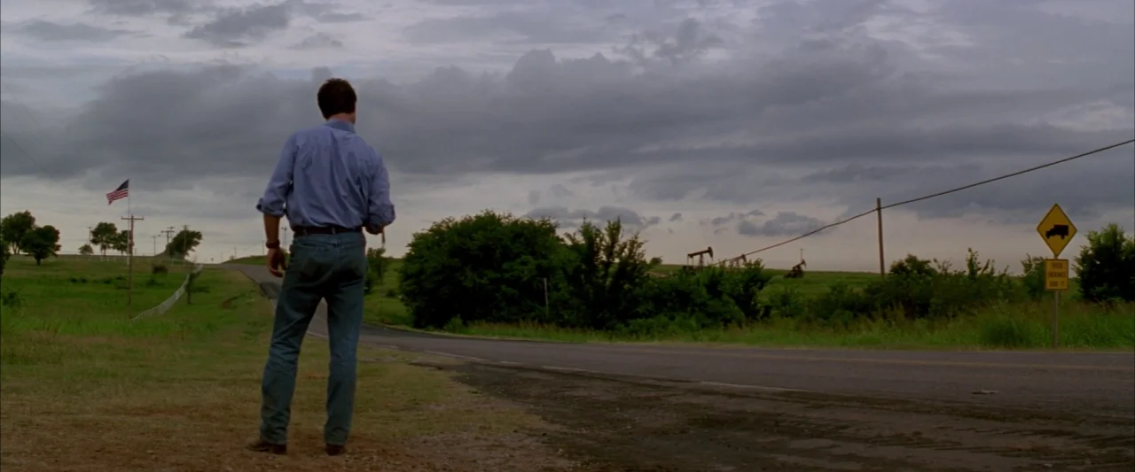 Twister (1996) movie still: fullbody — An asphalt road curves through a rural landscape under a heavily overcast, gray sky. A man in jeans and a…