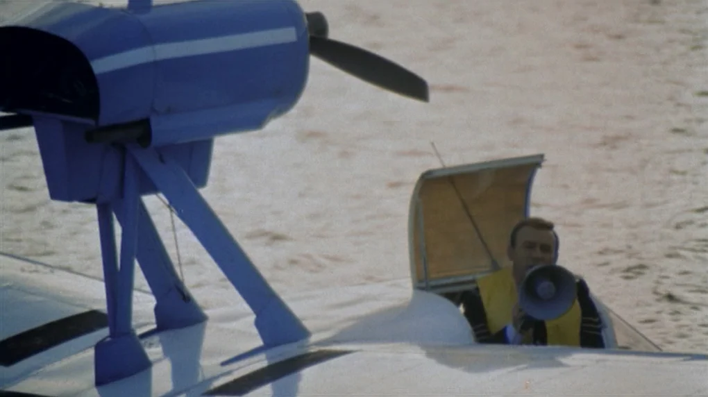 The Wicker Man movie still: wide — A blue and white seaplane floats on calm water during the day. A man sits in the open cockpit wearing a yellow…