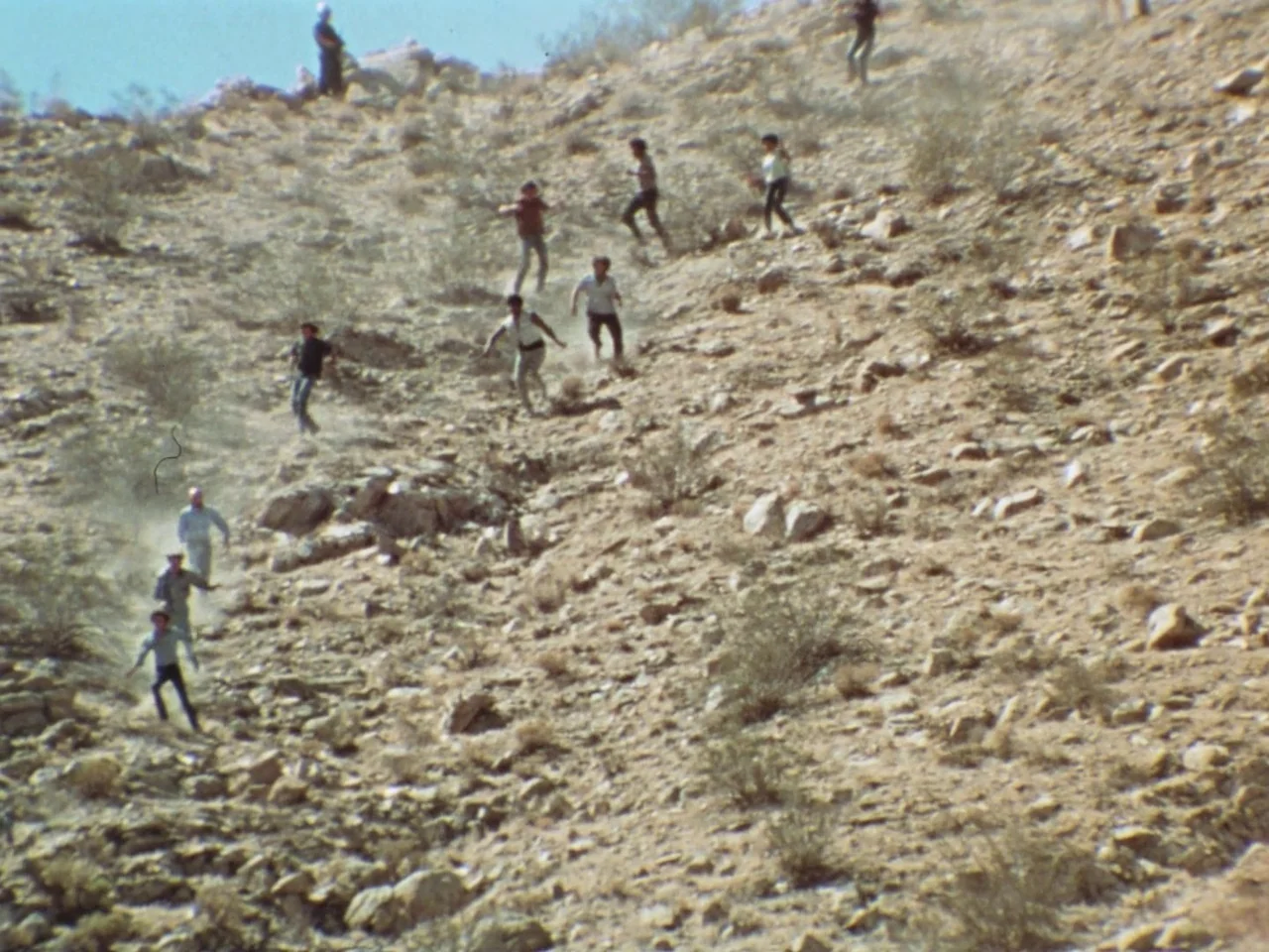 Punishment Park (1971) movie still: wide — A group of people navigate a steep, boulder-strewn hillside in a dry desert region. Multiple figures are…