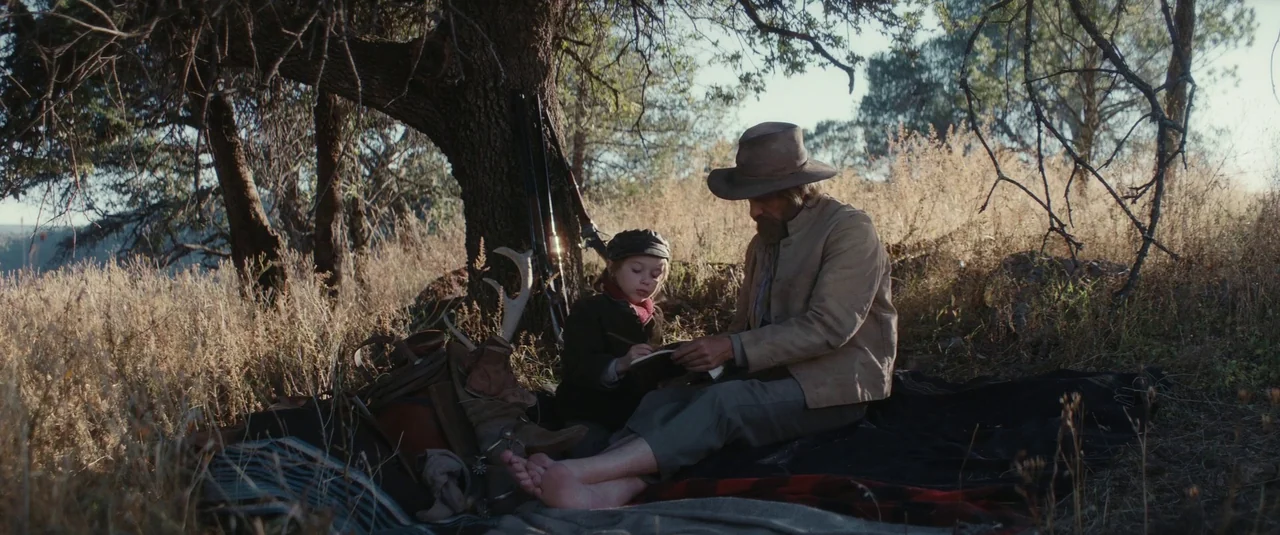 The Dead Don’t Hurt (2023) movie still: An older man and a child share a quiet, intimate moment of study on a remote, sun-bleached hillside. The…