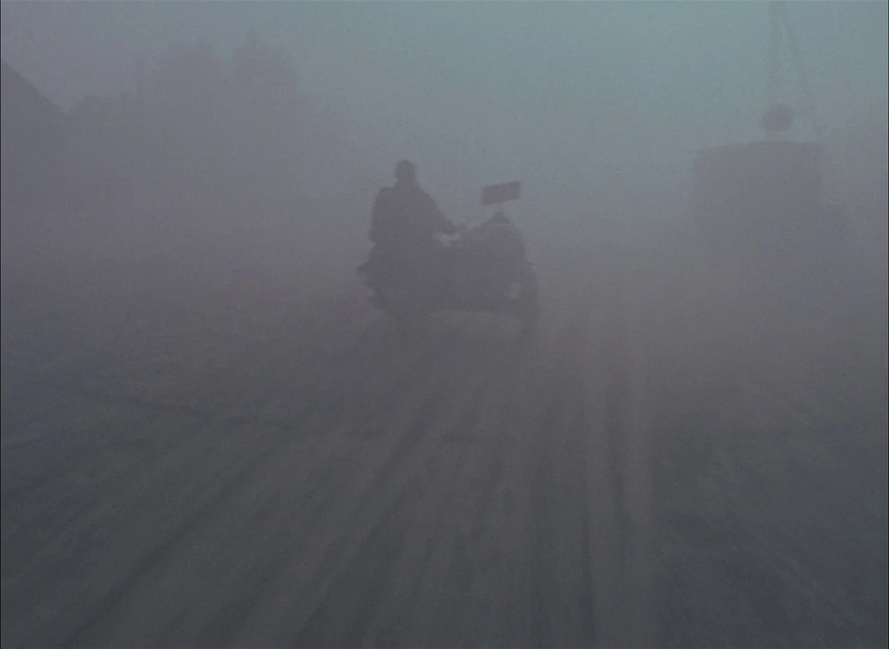 Come and See (1985) movie still: wide — An open road is completely obscured by dense, opaque fog in all directions. A lone motorcyclist drives away…