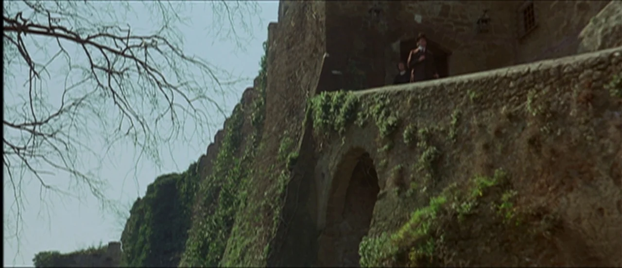 Flesh For Frankenstein (1973) movie still: wide — A stone castle wall features thick layers of ivy and a high stone archway. Two people stand near…