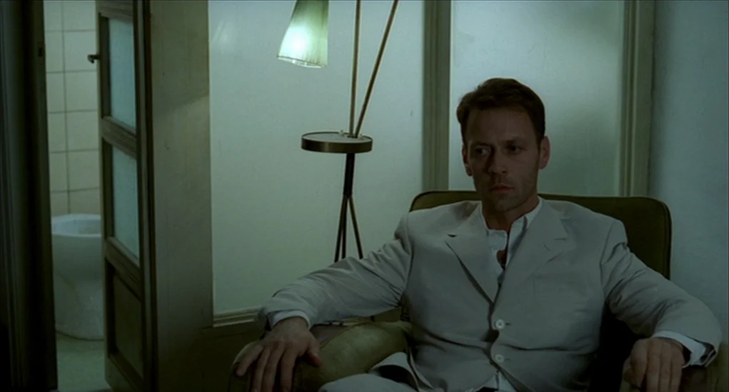 Anatomy of Hell (2004) movie still: medium — An interior space features a green chair and a floor lamp with a shade. The man sits in the chair while…