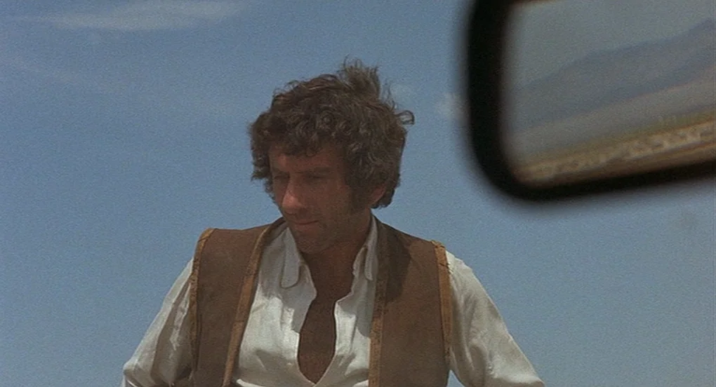 Vanishing Point (1971) movie still: medium — A man stands outdoors under a clear blue sky during the day. He looks down while wearing a white shirt…