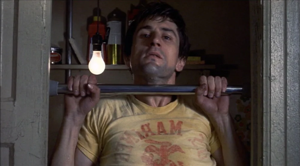 Taxi Driver (1976) movie still: medium — A dimly lit narrow doorway contains a metal pull-up bar fixed between wooden frames. A man in a yellow…