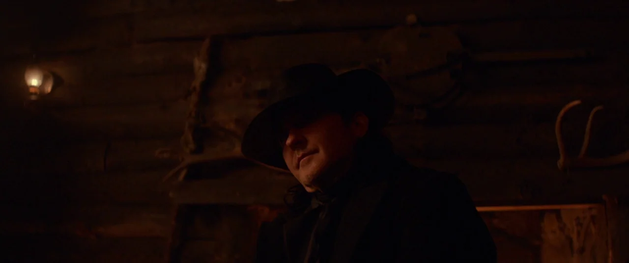 Never Grow Old (2019) movie still: medium — A dimly lit log cabin interior features wooden walls decorated with animal antlers. A man in a dark hat…