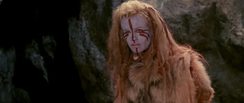 Clan of the Cave Bear (1986) movie still: medium — A dimly lit cave interior with textured rock walls behind the central figure. A woman with red…