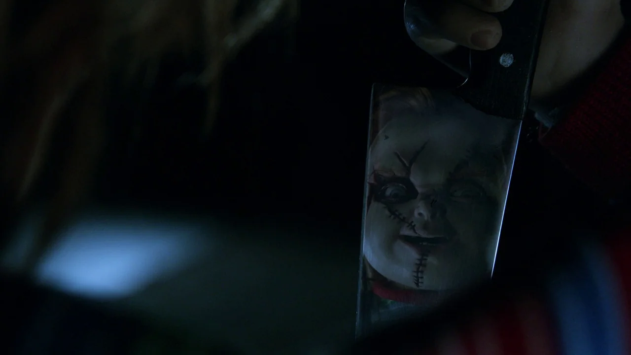 Curse of Chucky (2013) movie still: closeup — A dark space provides a black background for a held metal blade. The reflection of a scarred, stitched…
