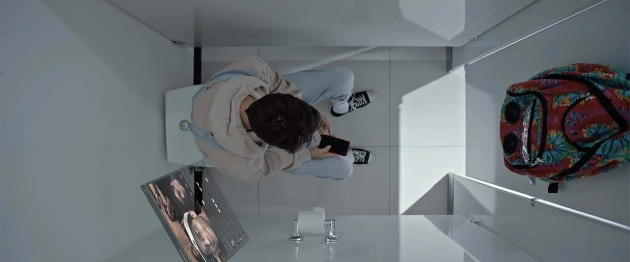 Init!ation (2021) movie still: wide — A stark white tiled bathroom stall contains a seated person and a colorful backpack. The person sits on the…
