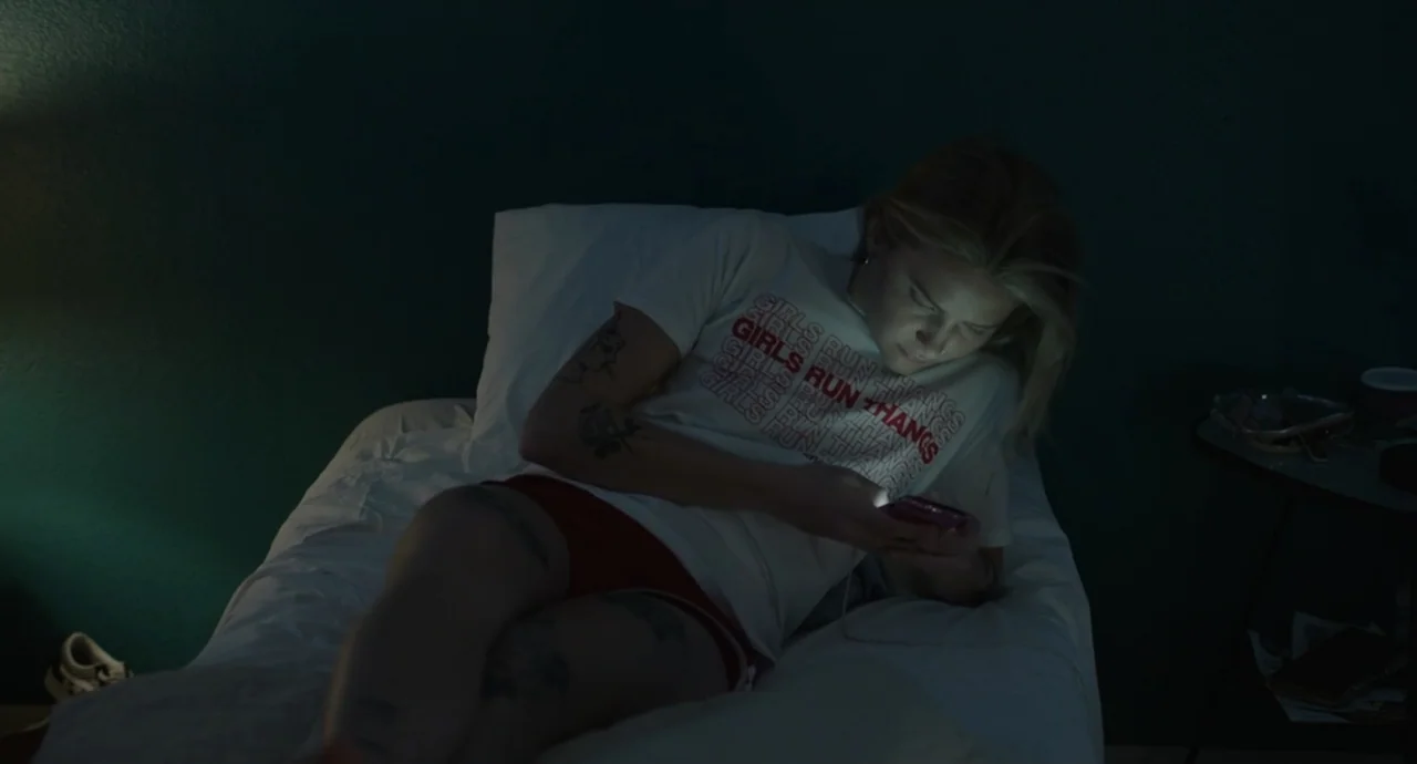 Pleasure (2021) movie still: medium — A dimly lit bedroom features a subject resting on a bed against dark green walls. The person holds a glowing…