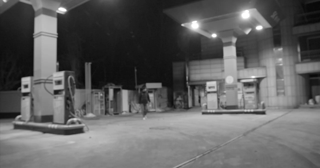 Hotel By The River movie still: wide — A deserted gas station at night illuminated by bright overhead canopy lights. A single figure walks across…