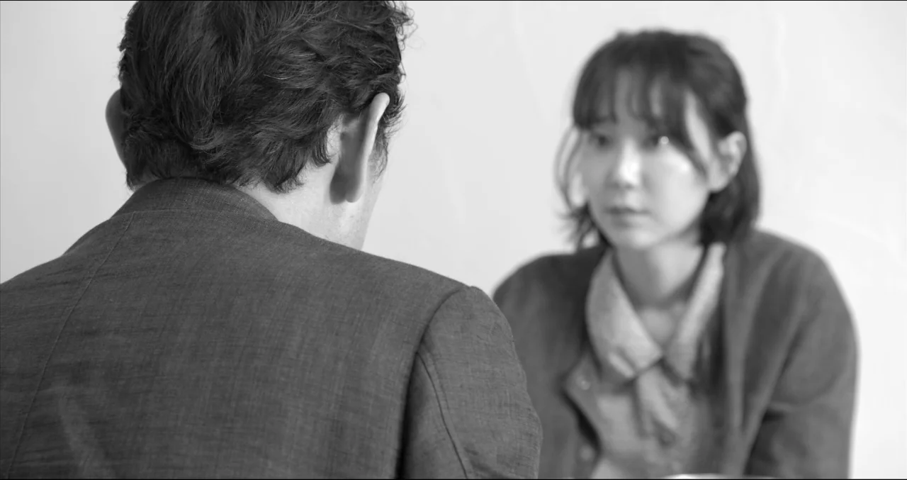 Grass (2018) movie still: medium — Two individuals sit in a brightly lit, minimalist space with featureless white walls. A man with his back to the…