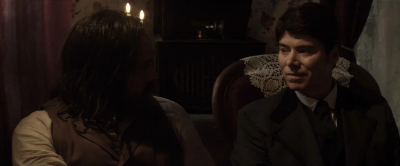 Matar o morir (1984) movie still: medium — A dimly lit 19th-century parlor with floral patterned wallpaper and antique furniture. Two men sit across…