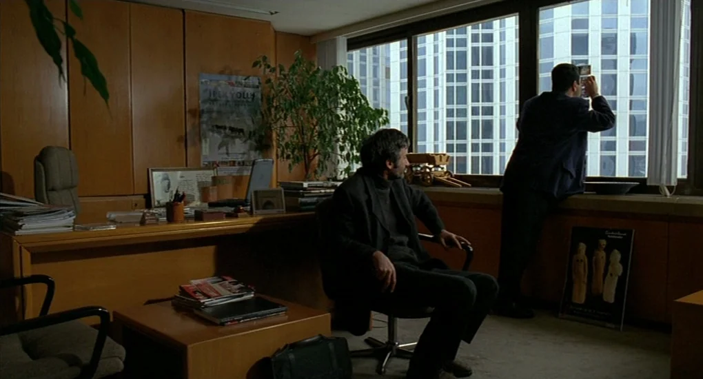 Uzak (2002) movie still: wide — An office features wood-paneled walls, a large desk, and floor-to-ceiling windows looking onto buildings. One man…