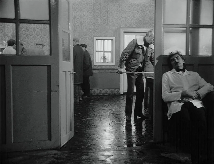 Damnation (1988) movie still: wide — A utilitarian room with patterned wallpaper and a reflective floor shows several men. A man wearing a white…