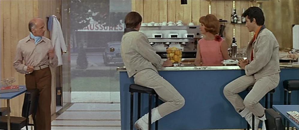 Les Demoiselles de Rochefort (1967) movie still: wide — A 1960s cafe with wood-paneled walls and a blue counter. Four people interact inside the…