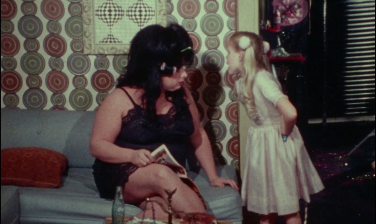 Female Trouble (1974) movie still: medium — A domestic room features circular-patterned wallpaper and a grey upholstered couch. A young girl stands…