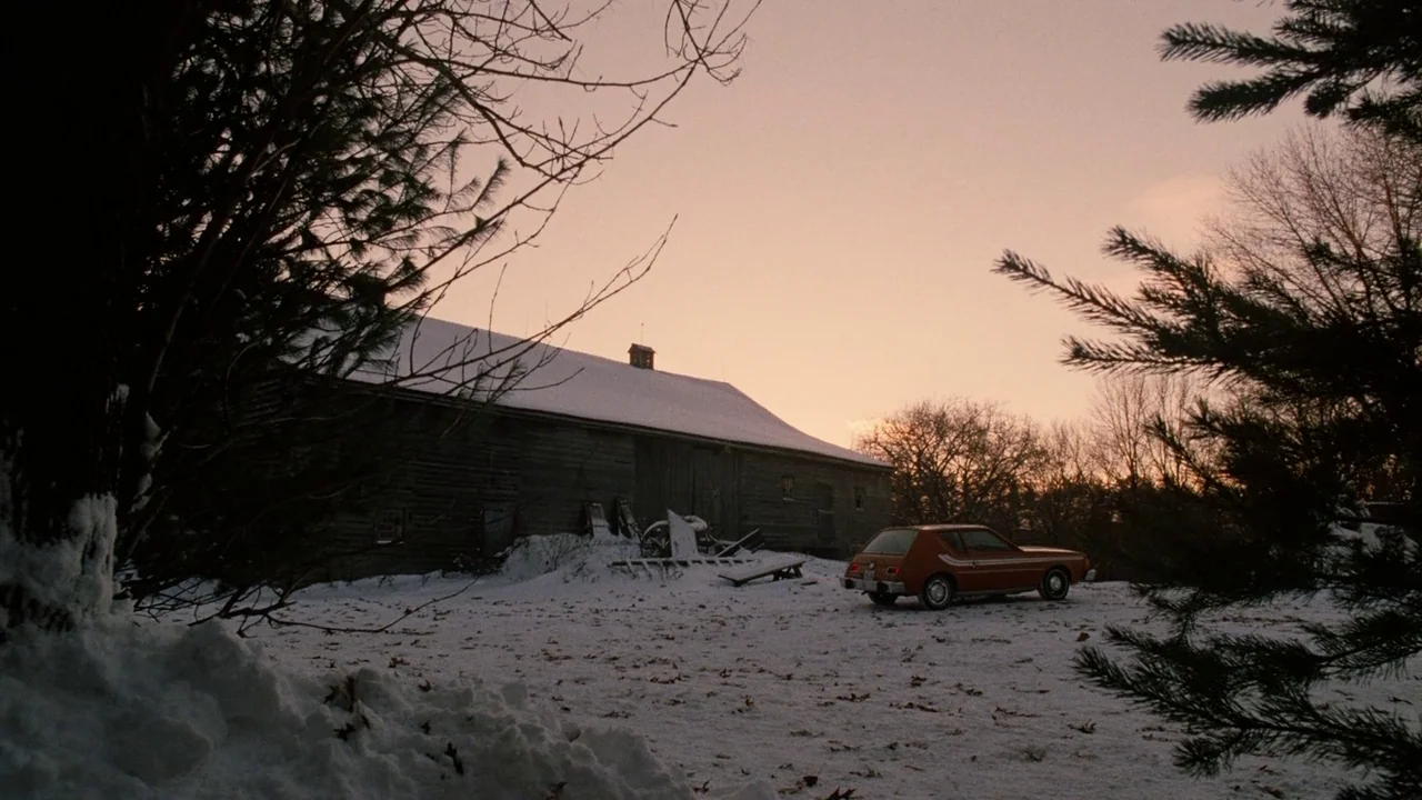 Splice (2009) movie still: wide — A weather-worn wooden barn stands in a snow-covered field under a pastel winter sky. A small orange car is parked…