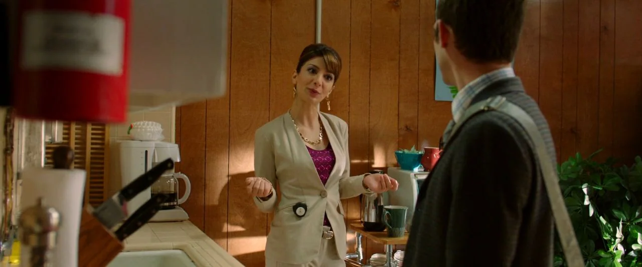 Cooties (2014) movie still: medium — An interior space with vertical wood-paneled walls and a tiled countertop area. A woman in a business suit…
