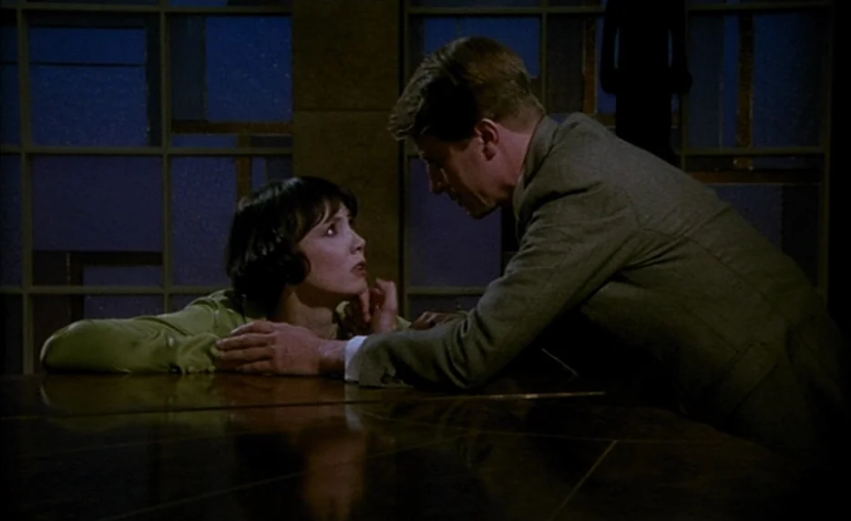 Mélo (1986) movie still: medium — A dimly lit room with large geometric glass panels creating a dark night backdrop. A man in a suit leans across a…