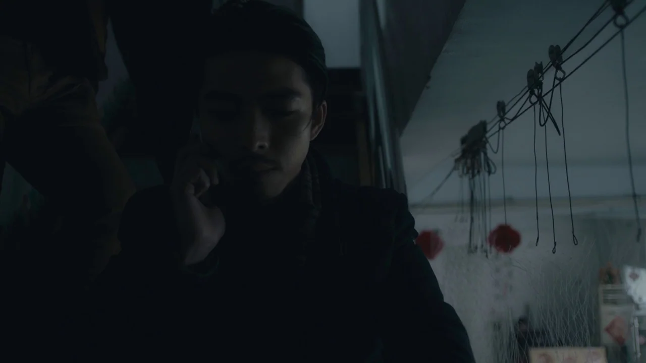 An Elephant Sitting Still (2018) movie still: medium — A dimly lit staircase features a man sitting quietly in the shadows. He holds a phone to his…