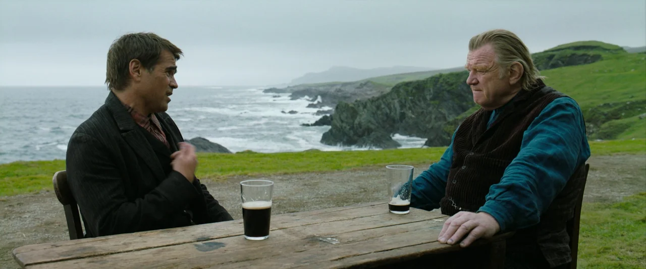 The Banshees of Inisherin (2022) movie still: medium — A wooden table sits on a grassy cliff overlooking a rocky coastline and ocean. Two men sit…