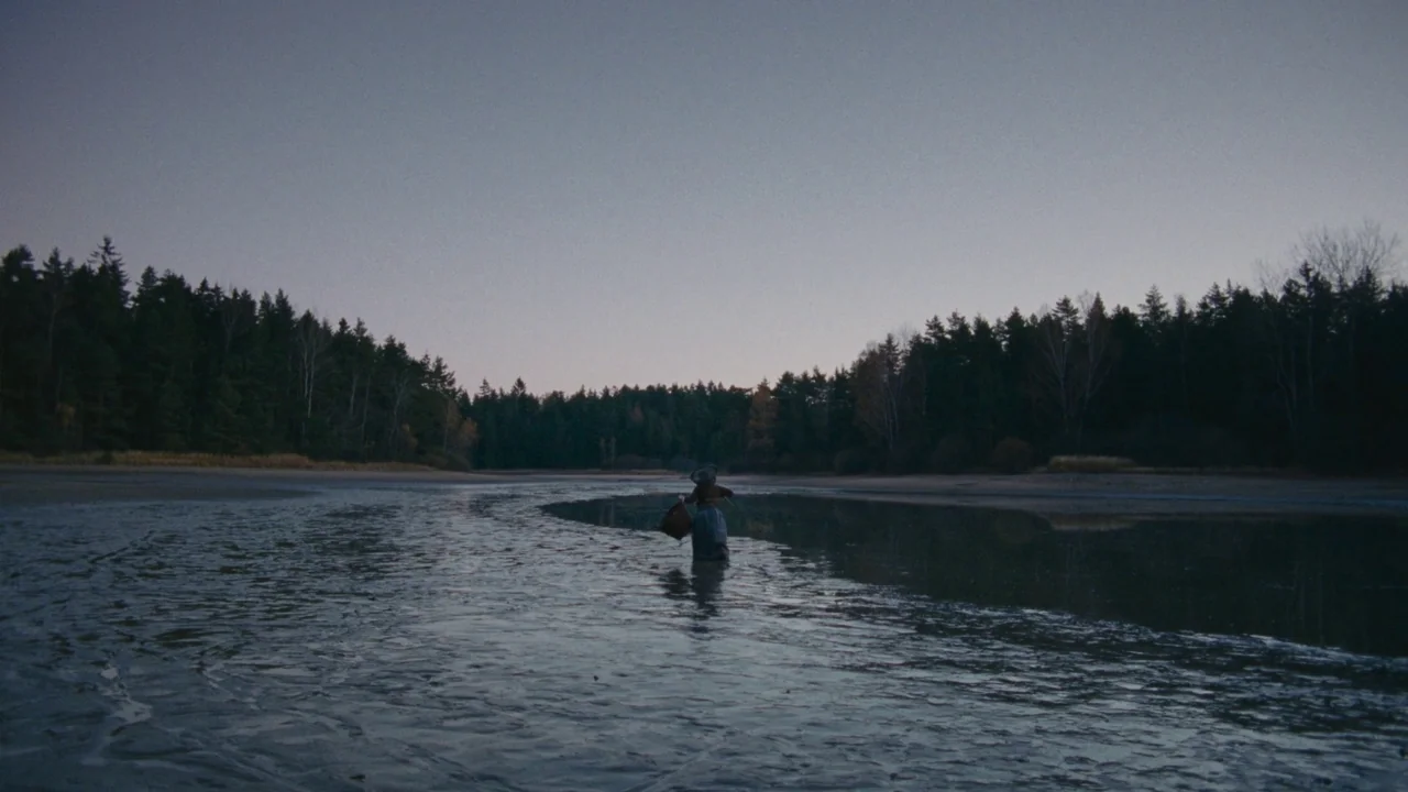 The Devil’s Bath (2024) movie still: wide — A wide, shallow river flows through a dense pine forest at twilight. A woman in a long dress walks…
