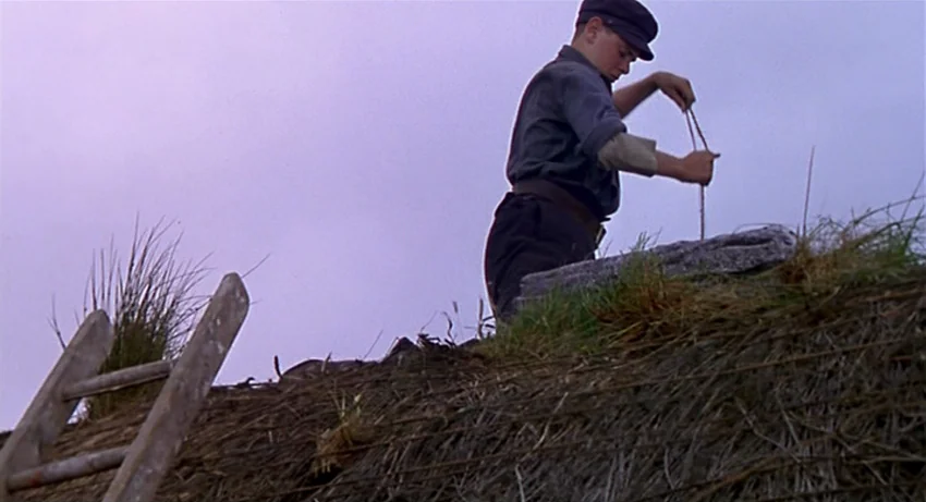 The Secret Of Roan Inish (1994) movie still: medium — A teenage boy works on top of a weathered, thick thatched roof. He pulls a thin rope taut…
