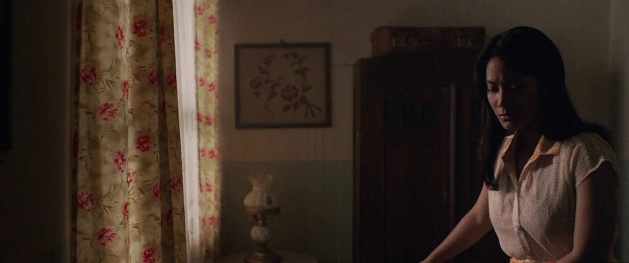 Satan’s Slaves (2017) movie still: medium — A dimly lit room with floral curtains and a wooden wardrobe against the wall. A woman with dark hair…