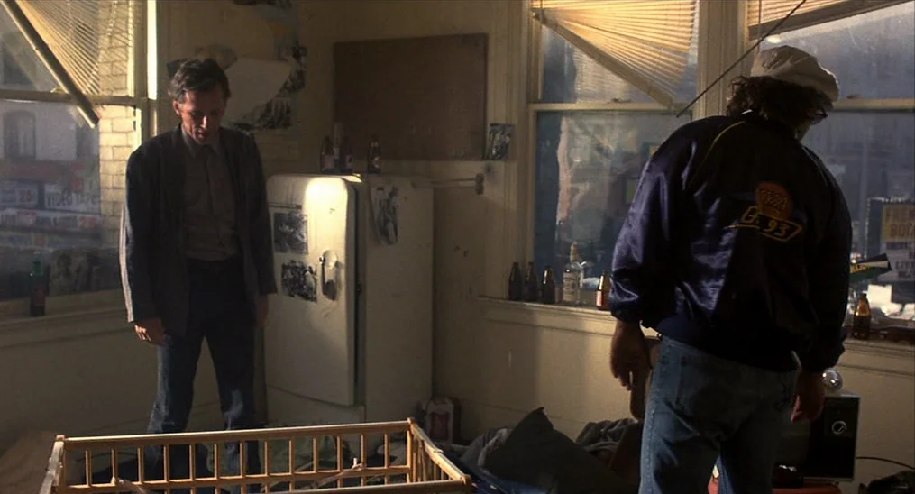 Salvador (1986) movie still: fullbody — An apartment interior features a white refrigerator and several beer bottles on windowsills. Two men stand…
