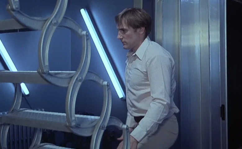 Maîtresse (1975) movie still: medium — A man stands in a sterile laboratory environment featuring dark walls and bright light fixtures. He gazes at…