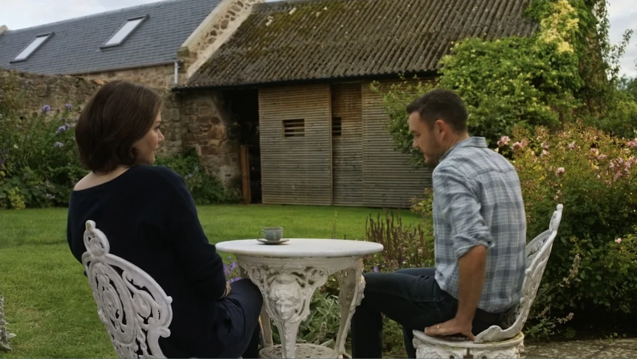 A Scottish Love Scheme (2024) movie still: medium — A man and woman sit at a white ornate metal table in a backyard. The background features a stone…