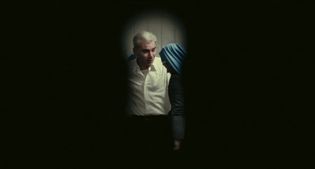 Todo Modo (1976) movie still: medium — A man in a white shirt interacts with a hooded figure in a dark area. The perspective mimics the view through…