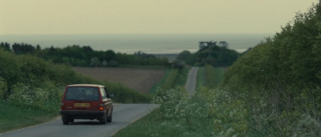 Never Let Me Go (2010) movie still: wide — A red station wagon travels along a narrow asphalt country road lined by thick hedges. In the distance, a…