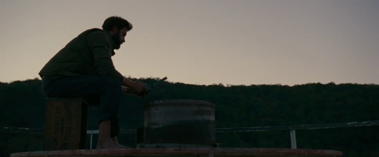 A Quiet Place (2018) movie still: wide — A man sits on a wooden crate on a dock at dusk. He looks down at a small object in his hands against a…