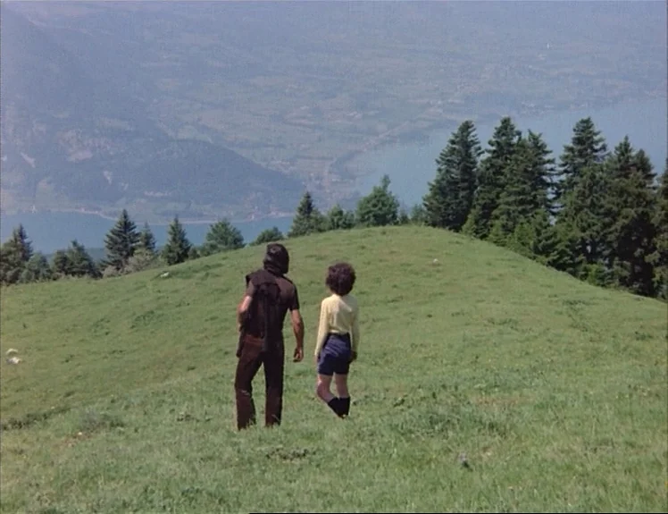 Claire’s Knee (1970) movie still: wide — A grassy mountainside overlooks a lake located in a distant valley below. Two figures stand together on the…